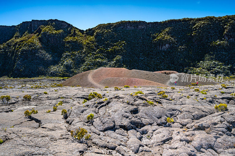 游客们走在天然火山景观中的小火山口前，火山口位于凝固的熔岩场中间，背景是一个巨大的绿色覆盖的火山口边缘。Formica Leo, Piton de la Fournaise，留尼旺岛，法国，欧洲。